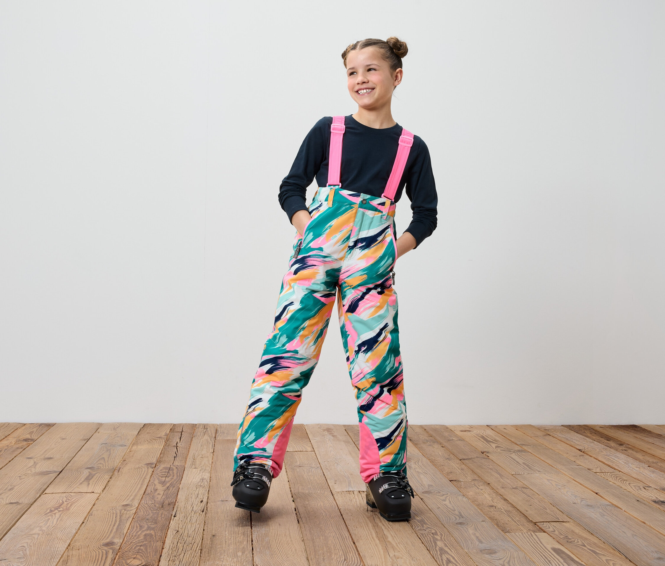 Usmívající se dospívající dívka v barevných lyžařských kalhotách s laclem a černých lyžařských botách stojí na dřevěné podlaze.