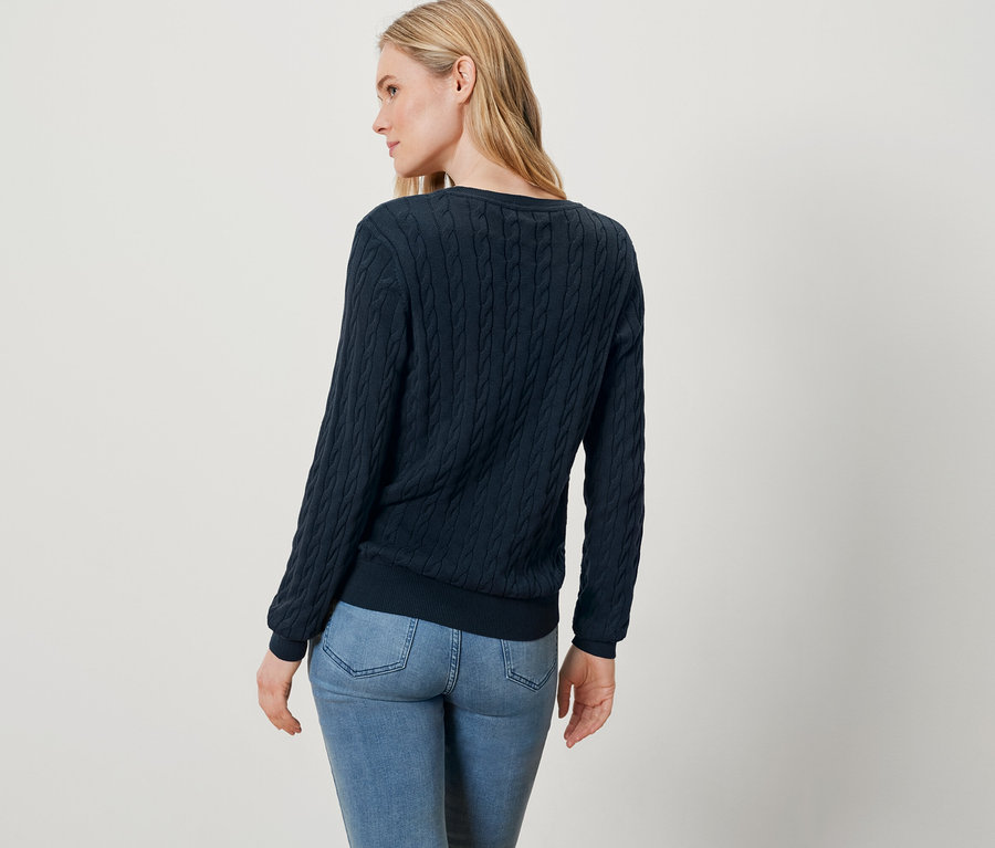 Žena s blond vlasy, oblečená v modrém svetru a džínách, stojí zády k fotoaparátu.