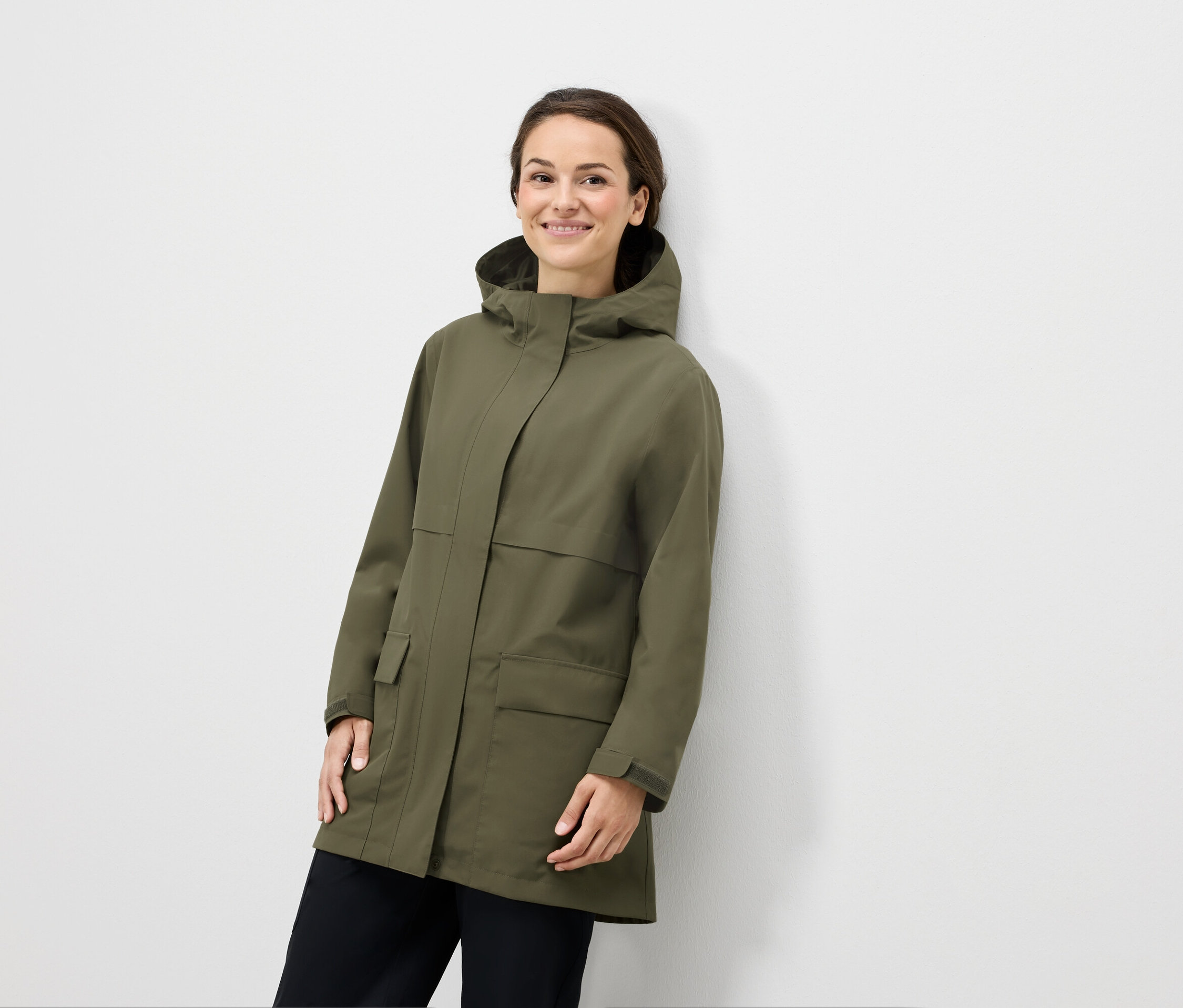 Žena pózuje v olivově zelené bundě s kapucí a černých kalhotách.