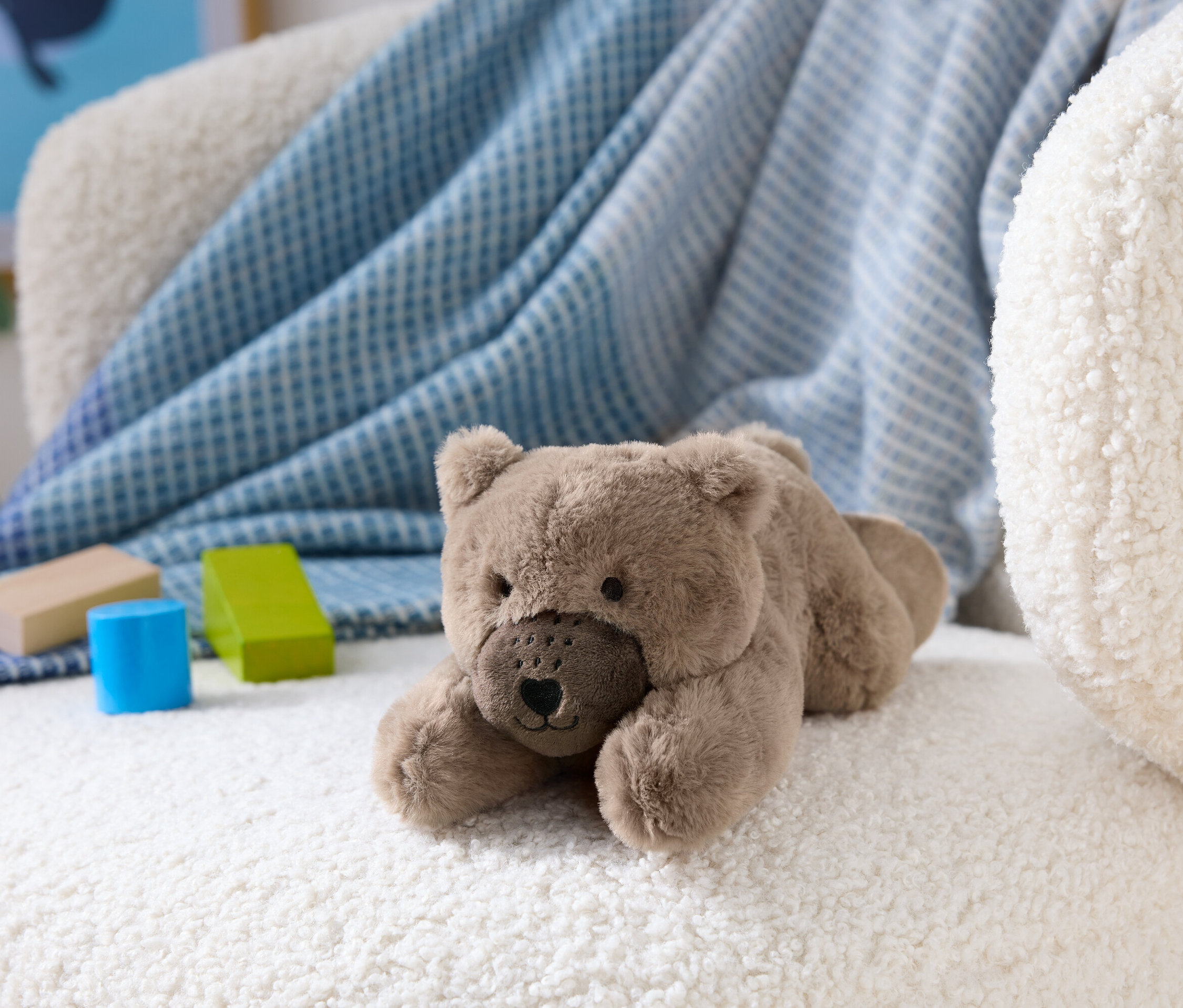 Hnědý plyšový medvěd leží na bílém huňatém křesle vedle dřevěných kostek.