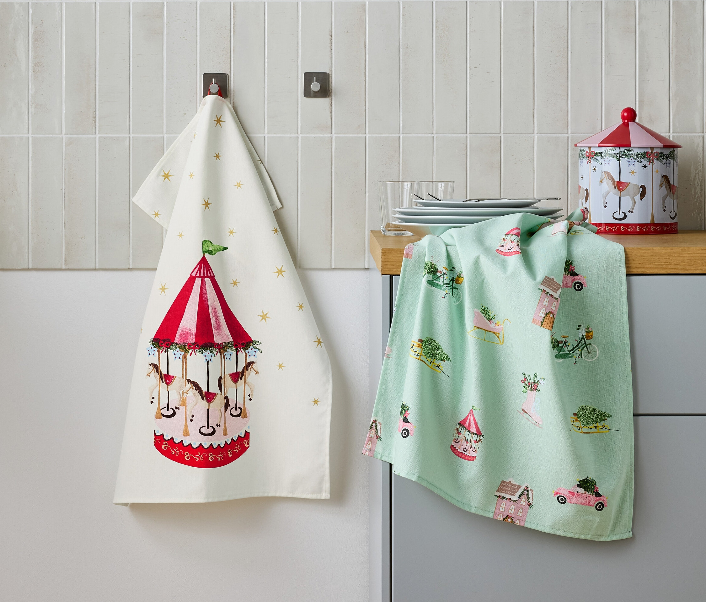 Dvě utěrky visí na zdi vedle sebe, jedna s motivem kolotoče a druhá se svátečními motivy, a plechová dóza na stole.