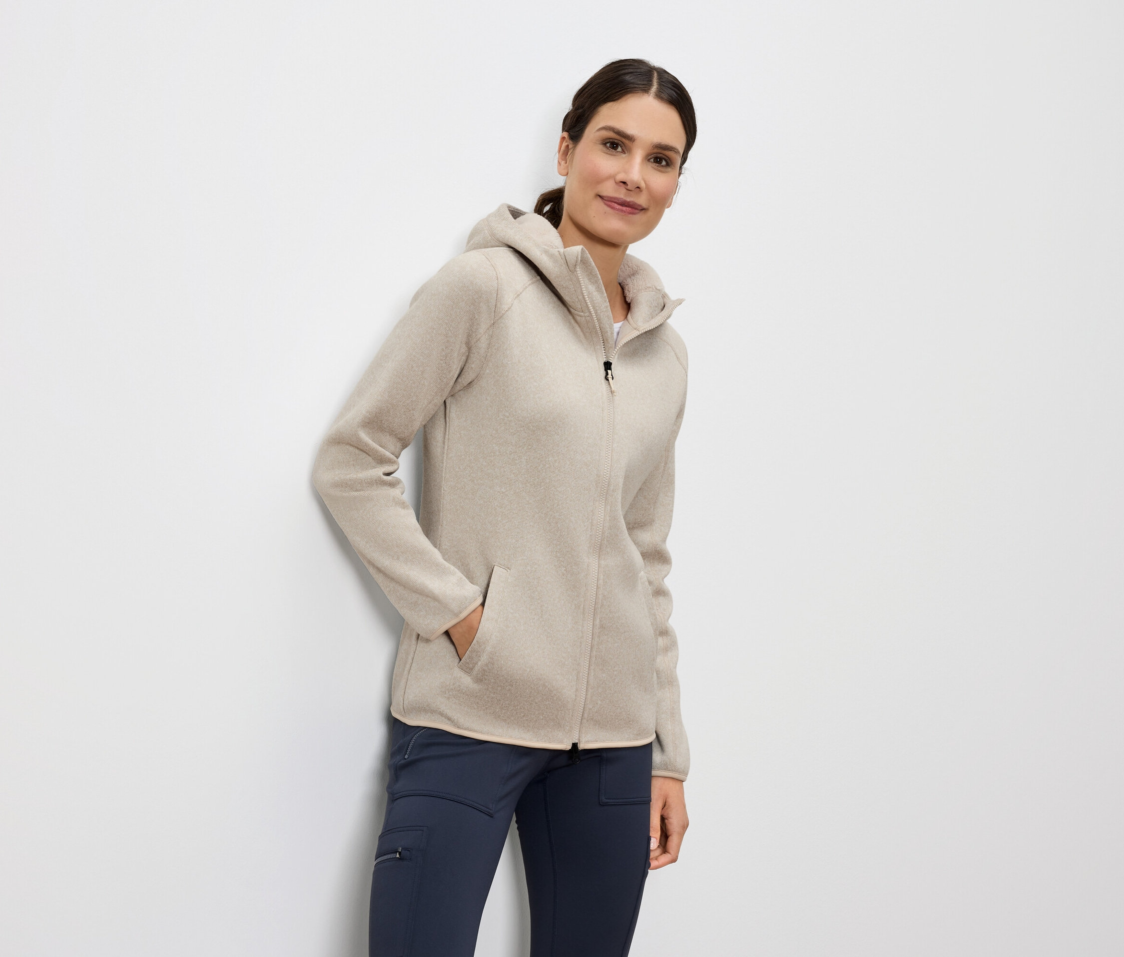 Žena stojí a má ruku v kapse béžové mikiny s kapucí na zip a modré kalhoty.