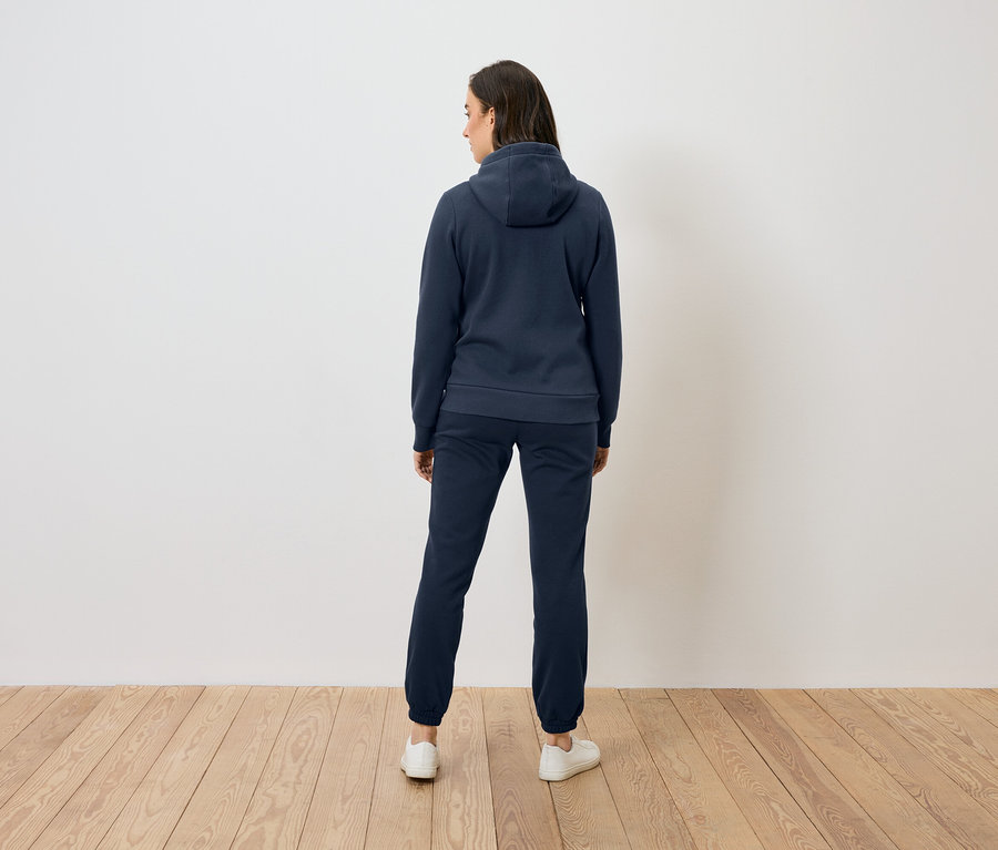 Žena zezadu v modré mikině s kapucí a kalhotách stojí na dřevěné podlaze před bílou zdí.