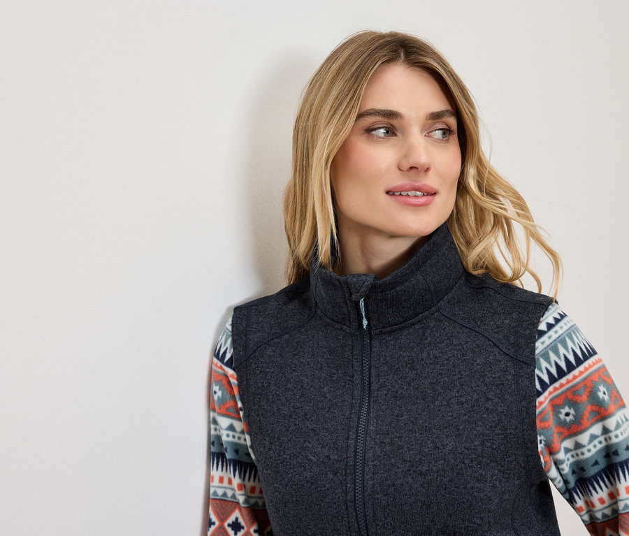 Blondýna v tmavě šedé vestě s ozdobným vzorem na rukávech se dívá doleva.