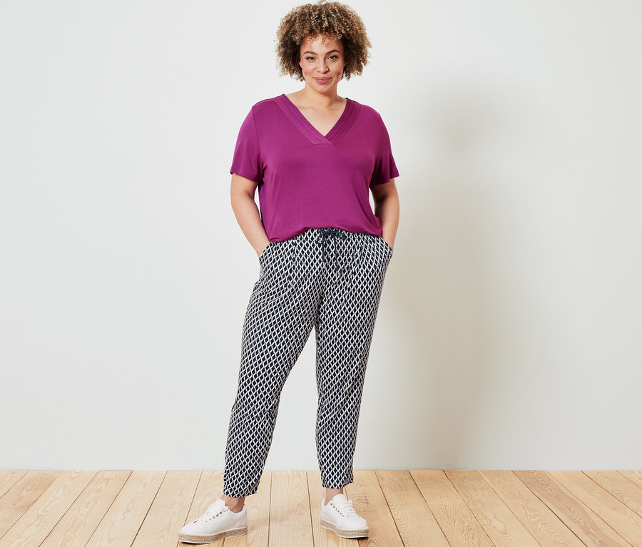 Žena pózuje v tričku s detailem skladů, bobulová a v žerzejových kalhotách s potiskem.