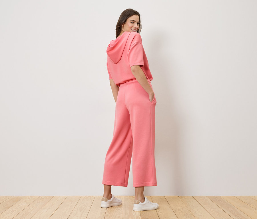 Žena se dívá přes rameno a má na sobě růžovou mikinu s kapucí a culottes kalhoty.