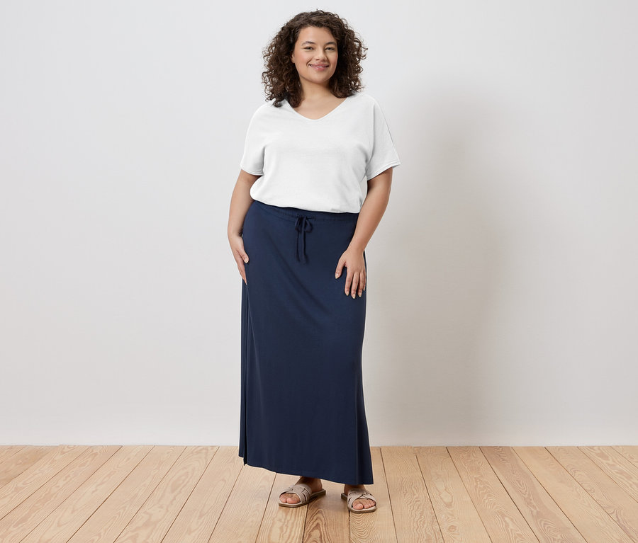 Žena stojí a má na sobě bílé tričko, dlouhou, tmavě modrou sukni a sandály.