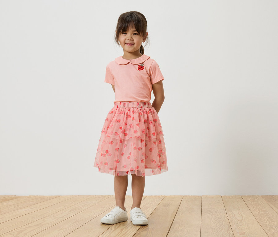 Dívka stojí před bílou zdí a má na sobě růžové tričko s jahodovým motivem, růžovou tylovou sukni s jahodami a bílé tenisky.