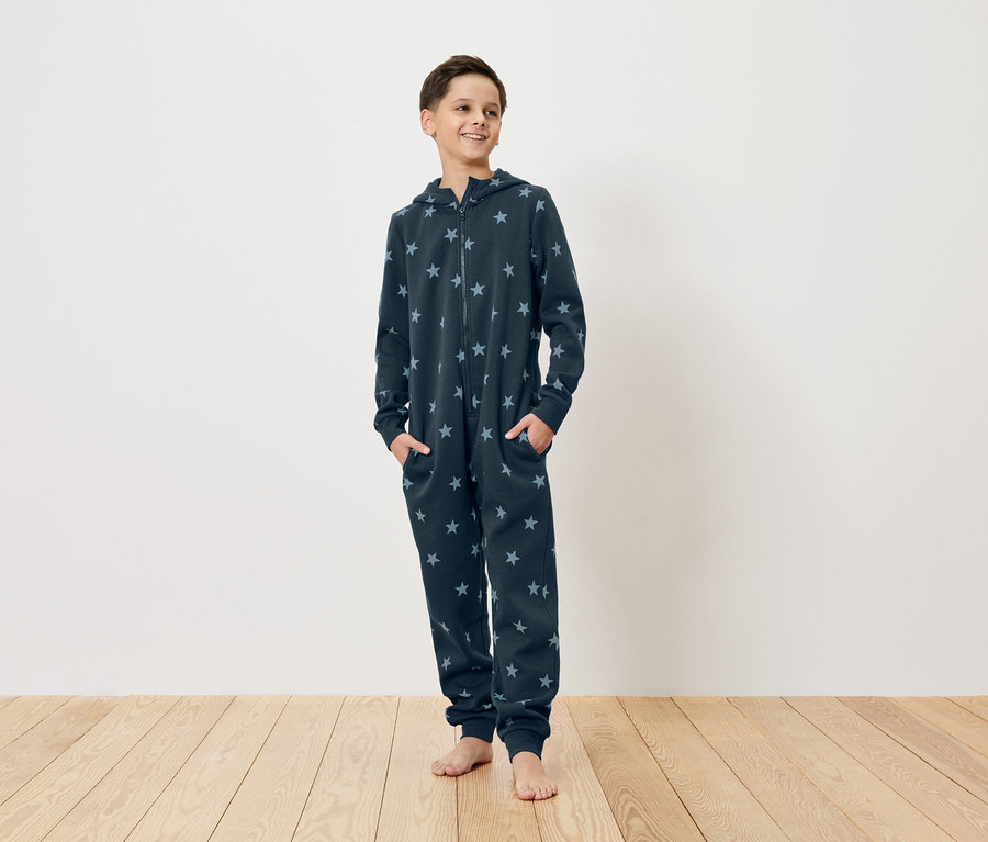Mladý muž stojí bosý na dřevěné podlaze, má na sobě tmavě modrý overal s hvězdami s kapucí a zipem a ruce v kapsách.