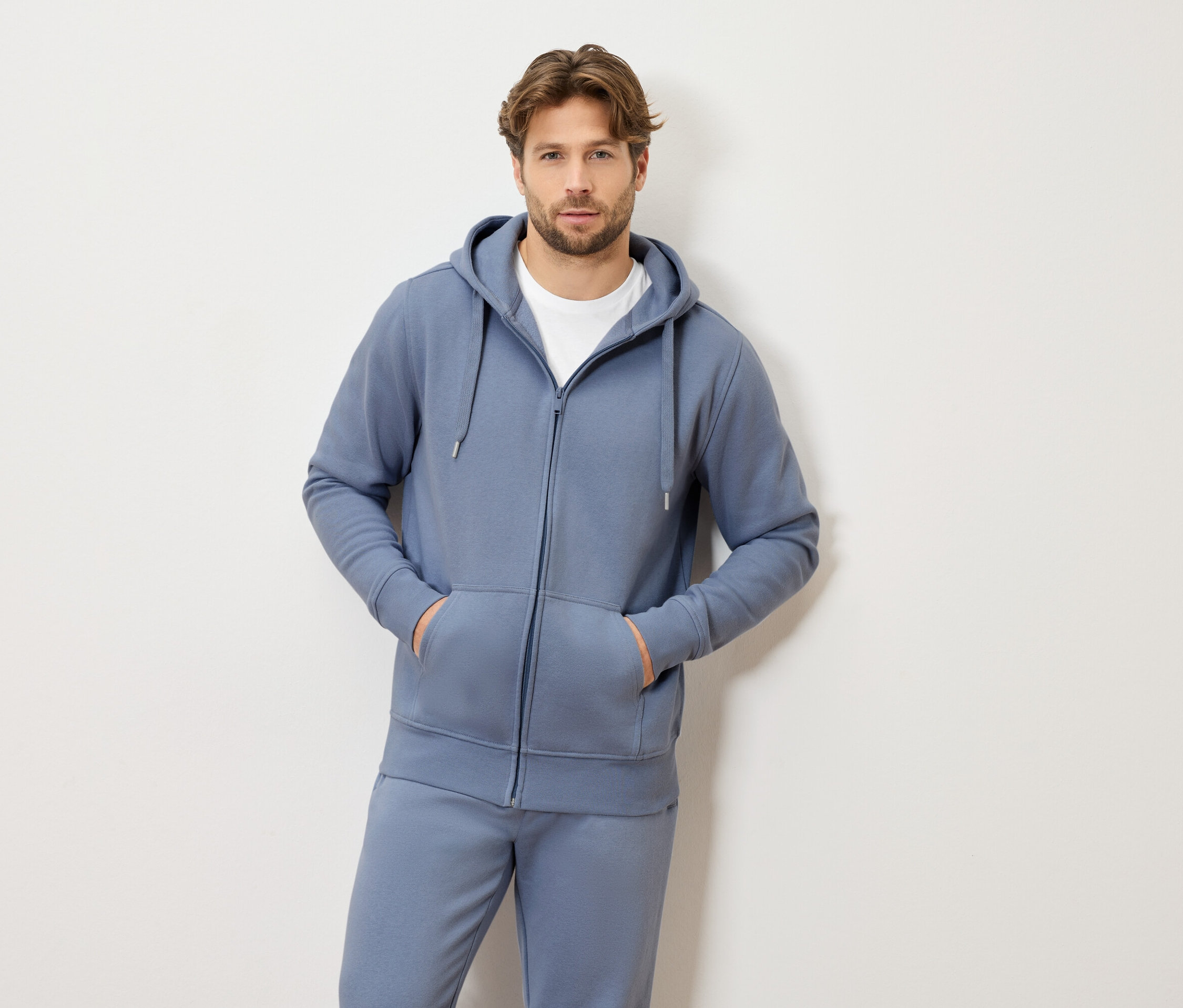 Muž se světle hnědými vlasy v modré teplákové soupravě s kapucí a zipem a bílém tričku stojí s oběma rukama v kapsách.