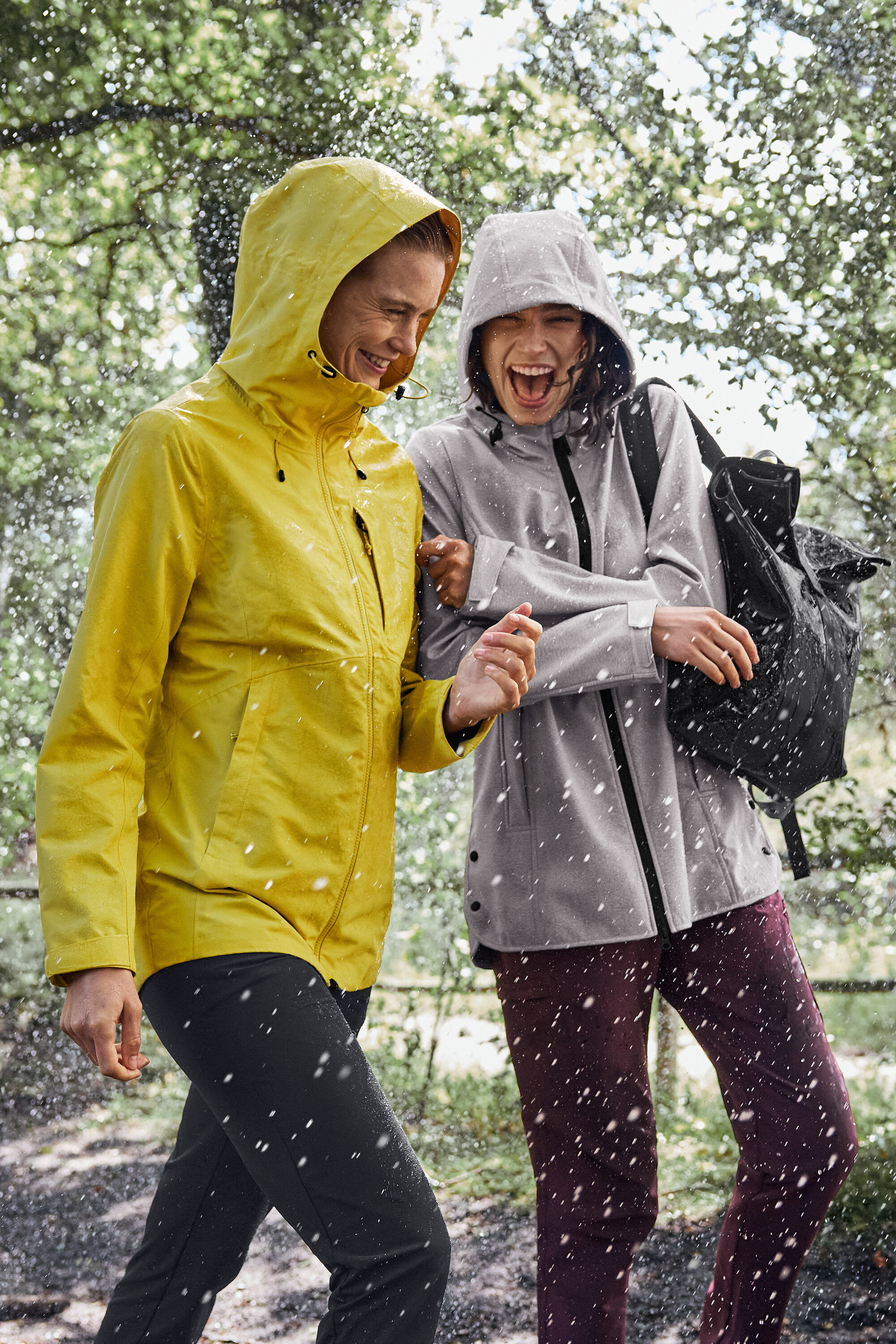 Dvě ženy jdou v dešti, jedna má na sobě žlutou pláštěnku, druhá šedou s černým batohem.