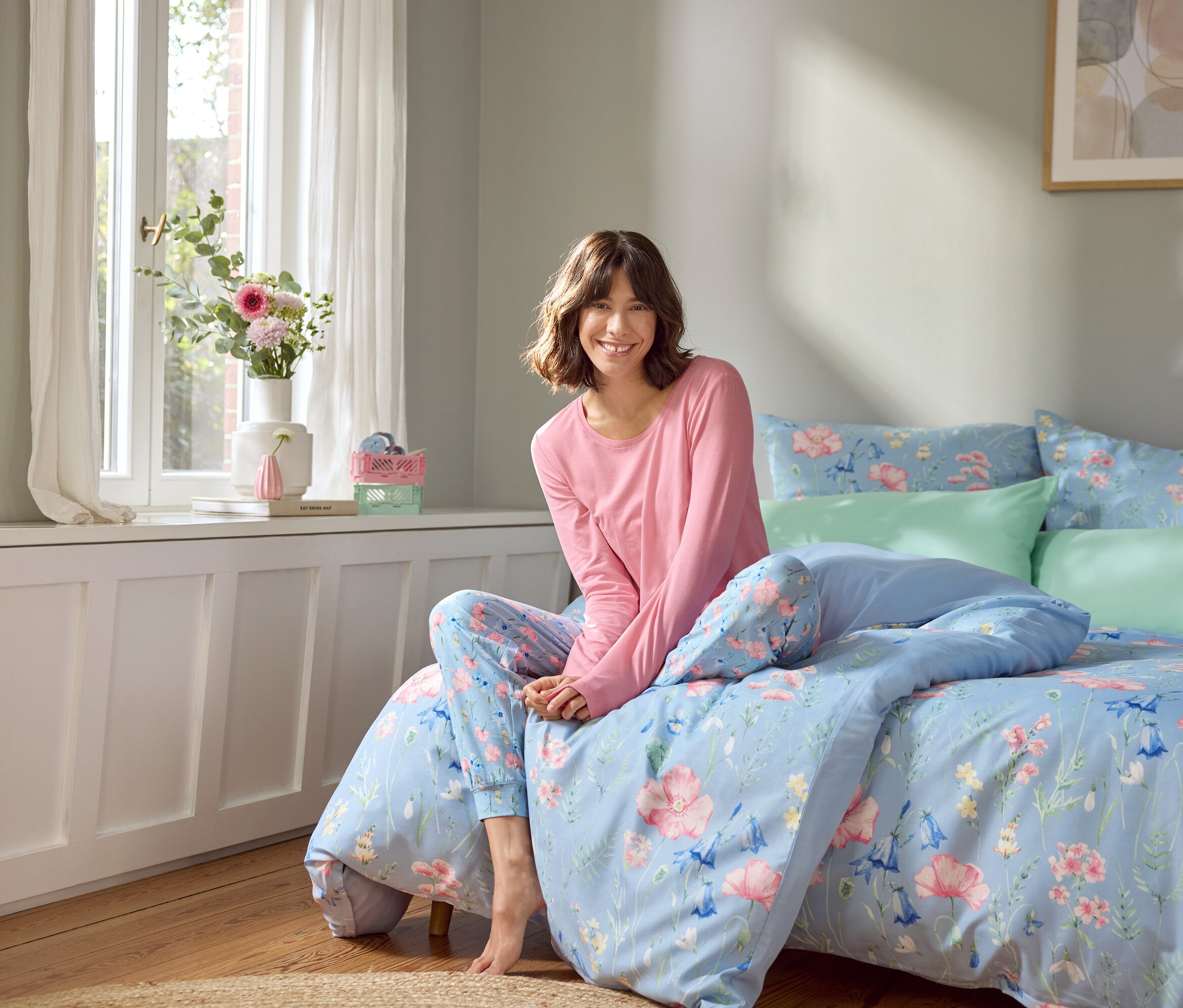 Žena sedí na posteli se světle modrým ložním prádlem s květinovým vzorem.