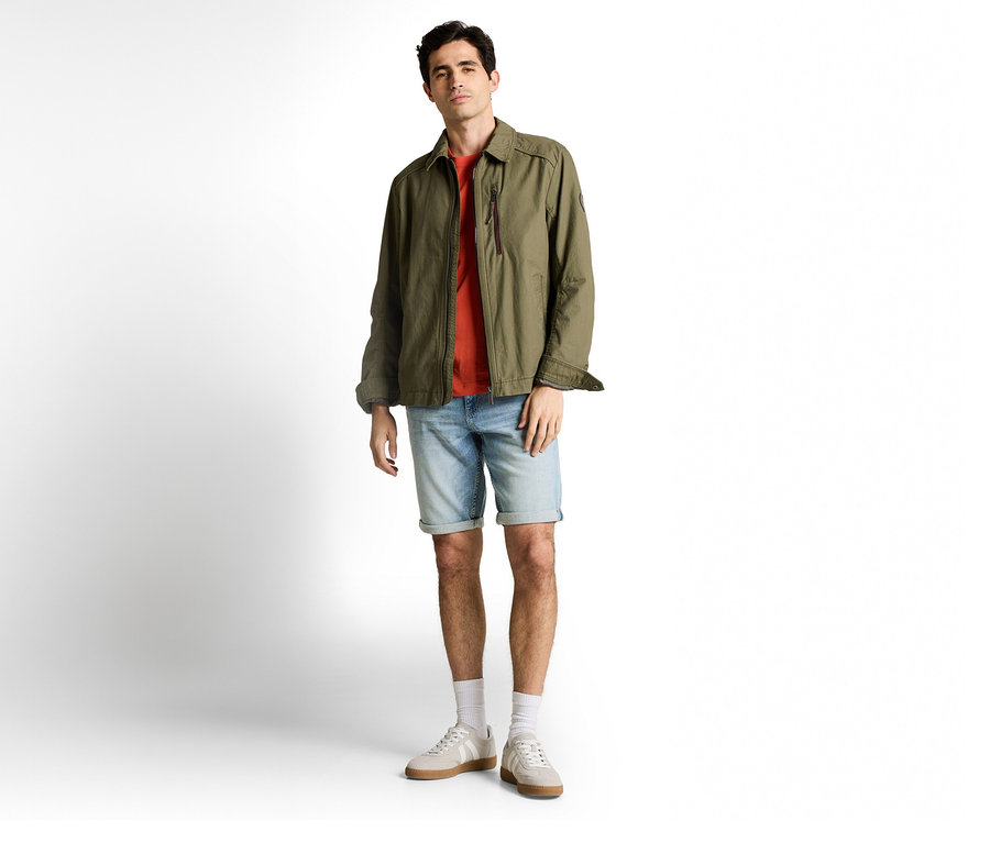 Muž v olivově zelené bundě a džínových šortkách Tom Tailor TTJOSH REGULAR SLIM, light stone blue denim, k tomu tričko, ponožky a tenisky.