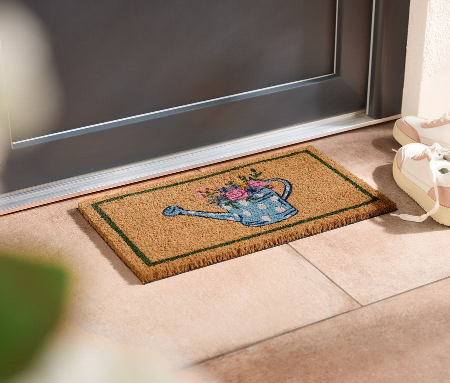 Rohožka s motivem konve leží před dveřmi, vedle stojí bílé tenisky.