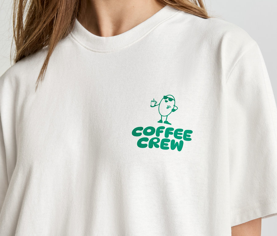 Detail osoby v světlém tričku se zeleným potiskem "Coffee Crew" a kreslenou postavičkou kávového zrna.