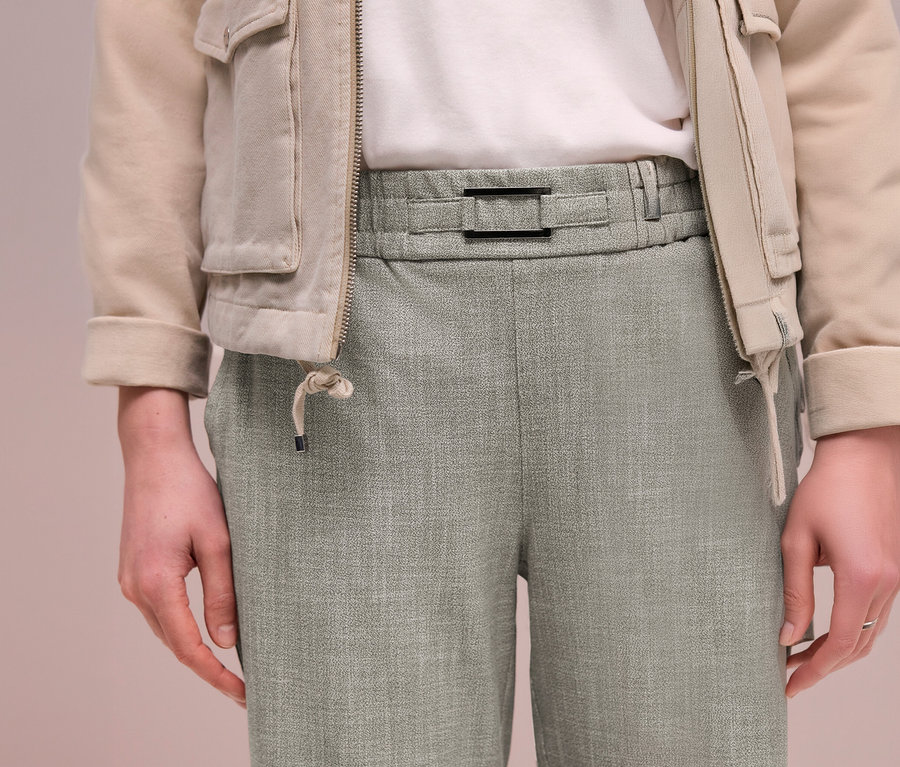 Osoba v béžové bundě, bílém tričku a světle zelených kalhotách joggpant Street One s černou sponou v pase.