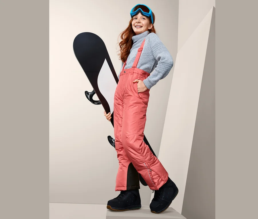 Usmívající se dívka drží snowboard a má na sobě lyžařské kalhoty, svetr a lyžařské brýle.