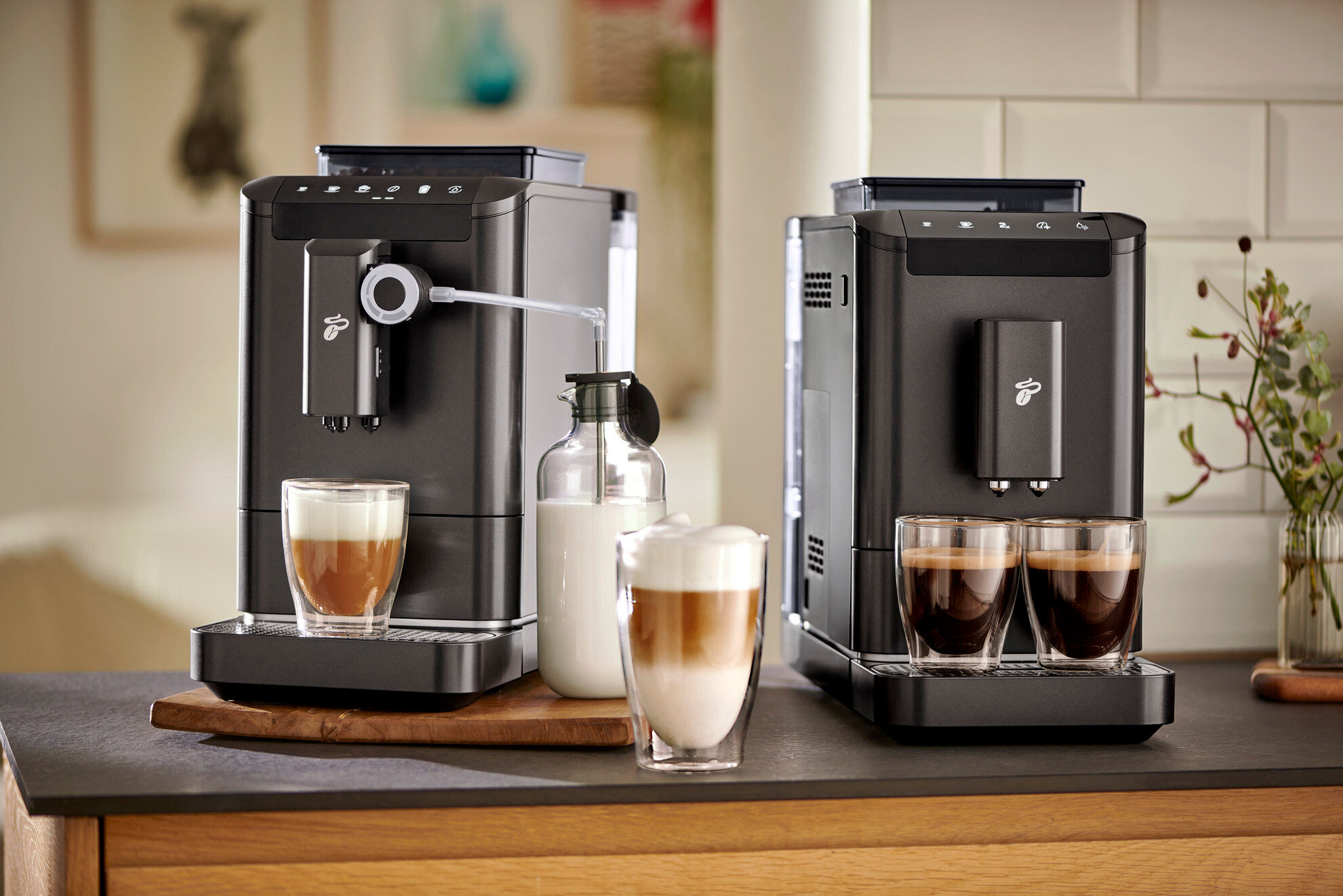 Dva černé kávovary stojí vedle sebe na desce stolu, před nimi jsou sklenice s kávou a mlékem.