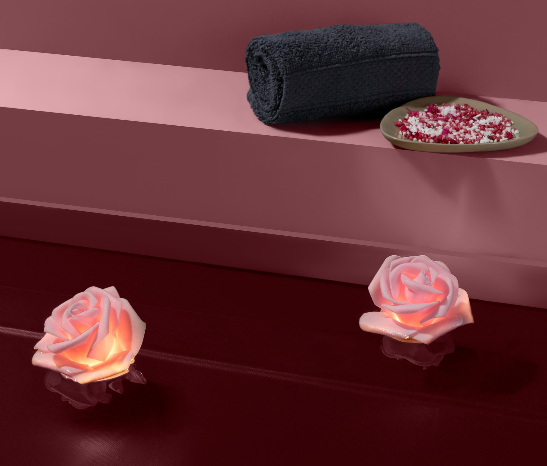 Dvě plovoucí světýlka s LED ve vzhledu růže na hladině vody. V pozadí je stočený ručník a miska s dekorativními kamínky.