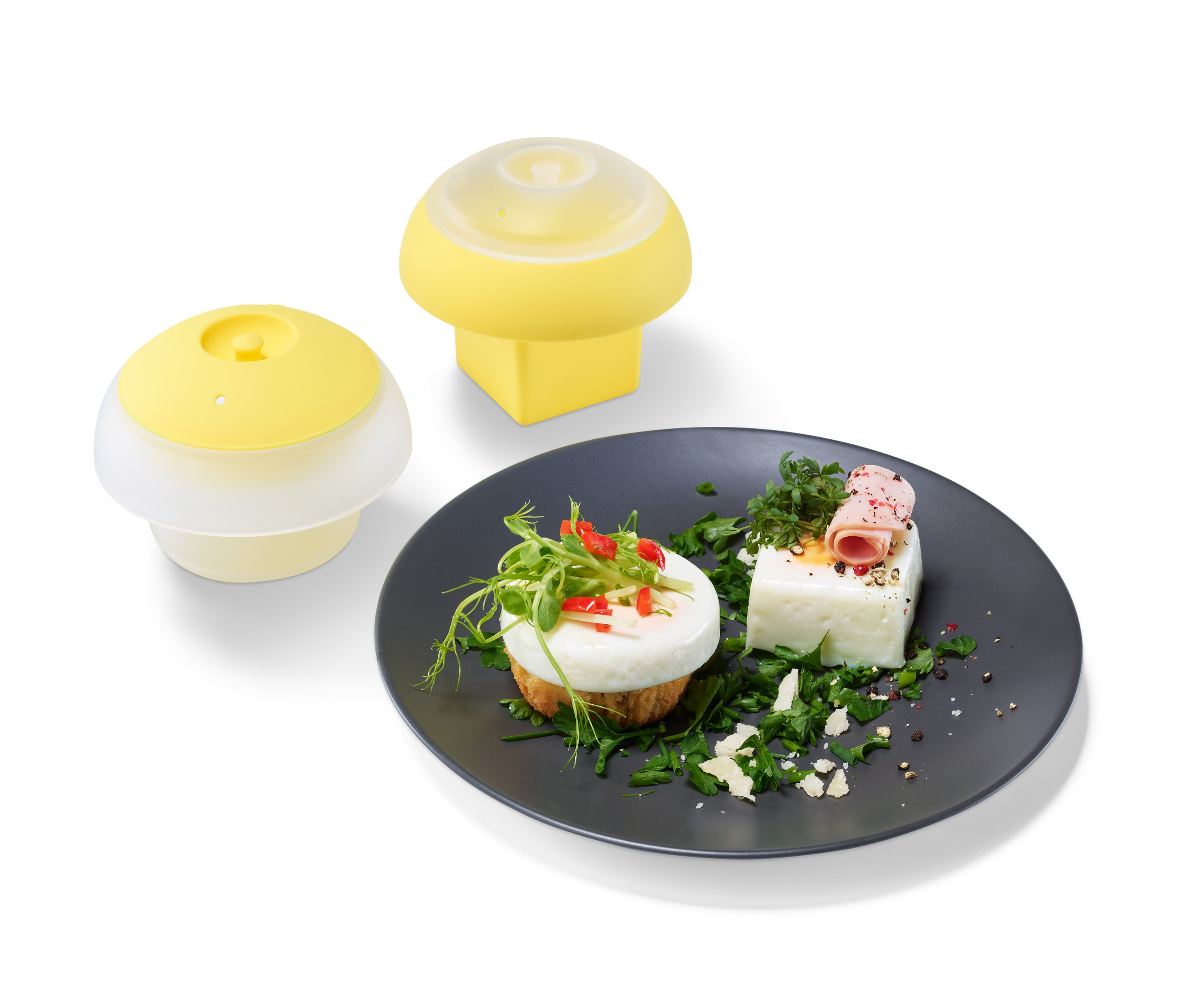 Dvě žluté formičky na vaření vajec a talíř s vejci zdobenými zelení.