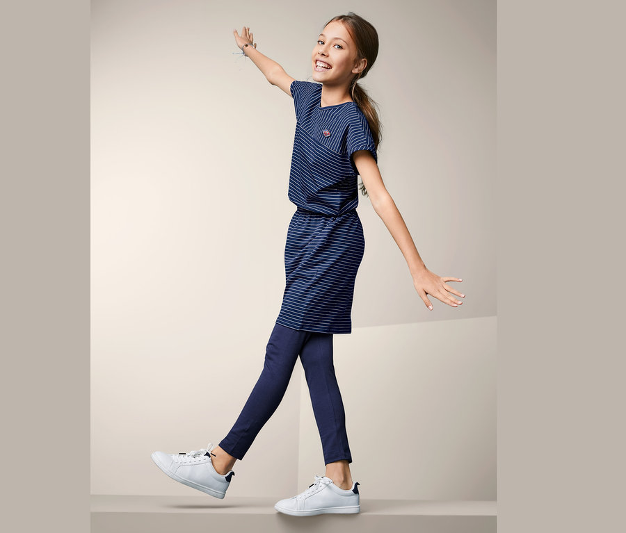 Usmívající se dívka balancuje v džerzejových šatech s modrými proužky, tmavě modrých legínách a bílých teniskách.