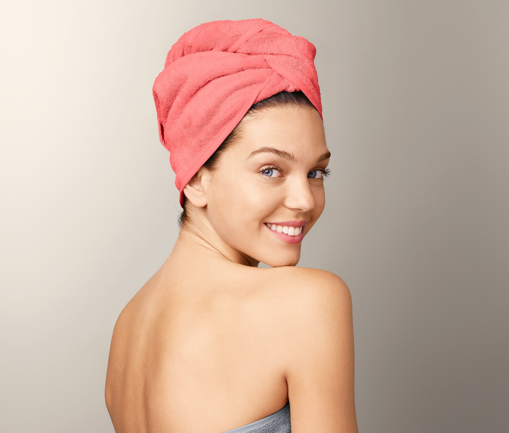 Žena se usmívá přes rameno, má na sobě ručníkový turban a je zabalená do šedé osušky.