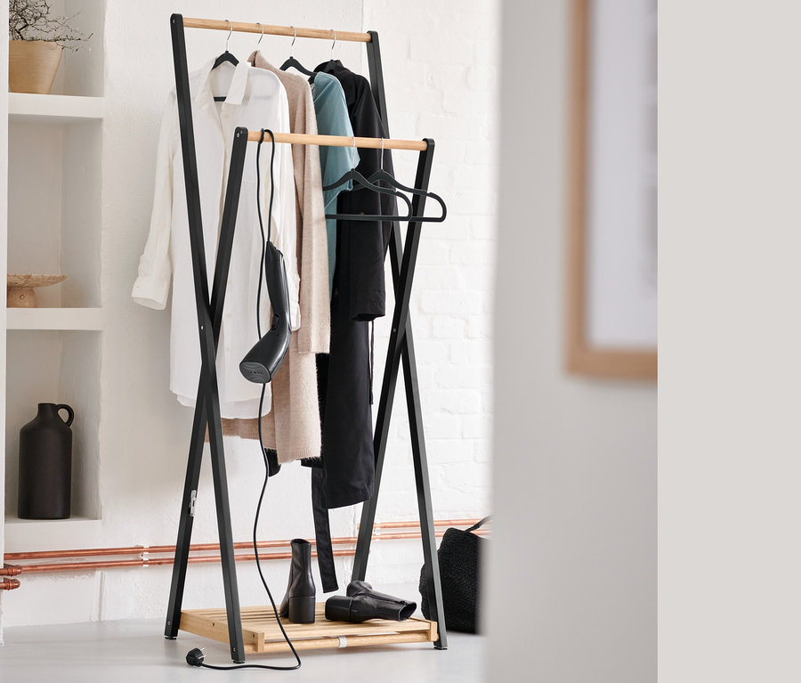 Oblečení visí na věšáku, pod ním stojí napařovač a černé ramínka na šaty.