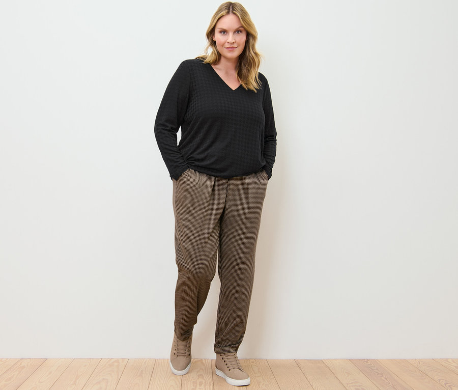 Žena stojí a má ruce v kapsách kalhot s žakárovým vzorem a černého strukturovaného trika s dlouhým rukávem.