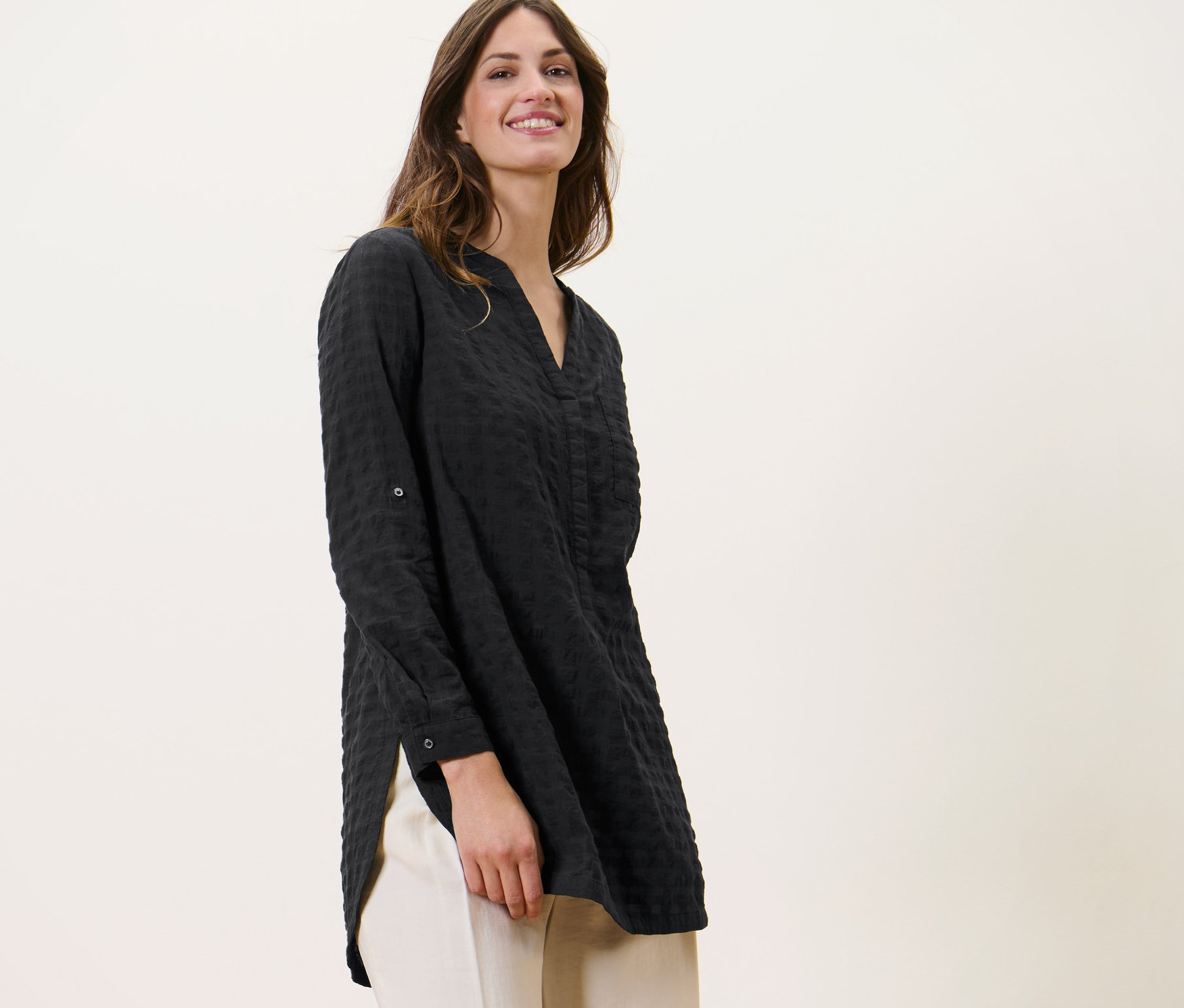 Žena má na sobě dlouhou tuniku s tkanou strukturou a světlé kalhoty.