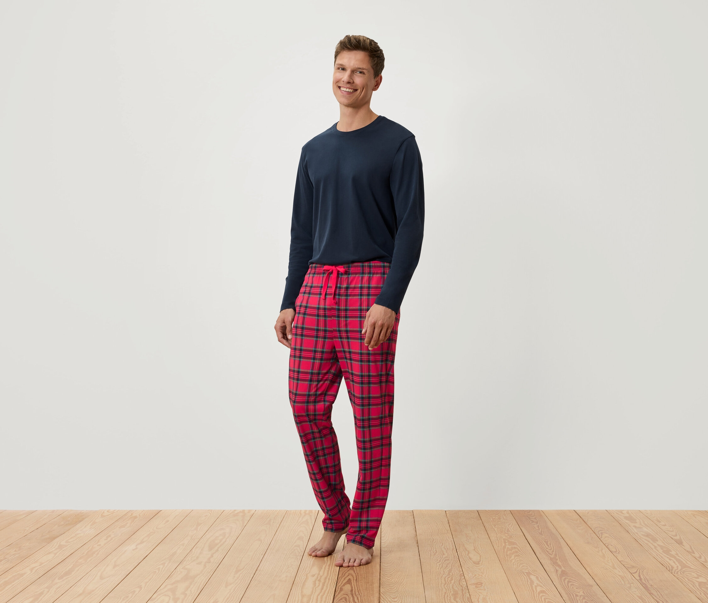 Muž stojí a má na sobě modré tričko s dlouhým rukávem a červené kostkované pyžamové kalhoty.