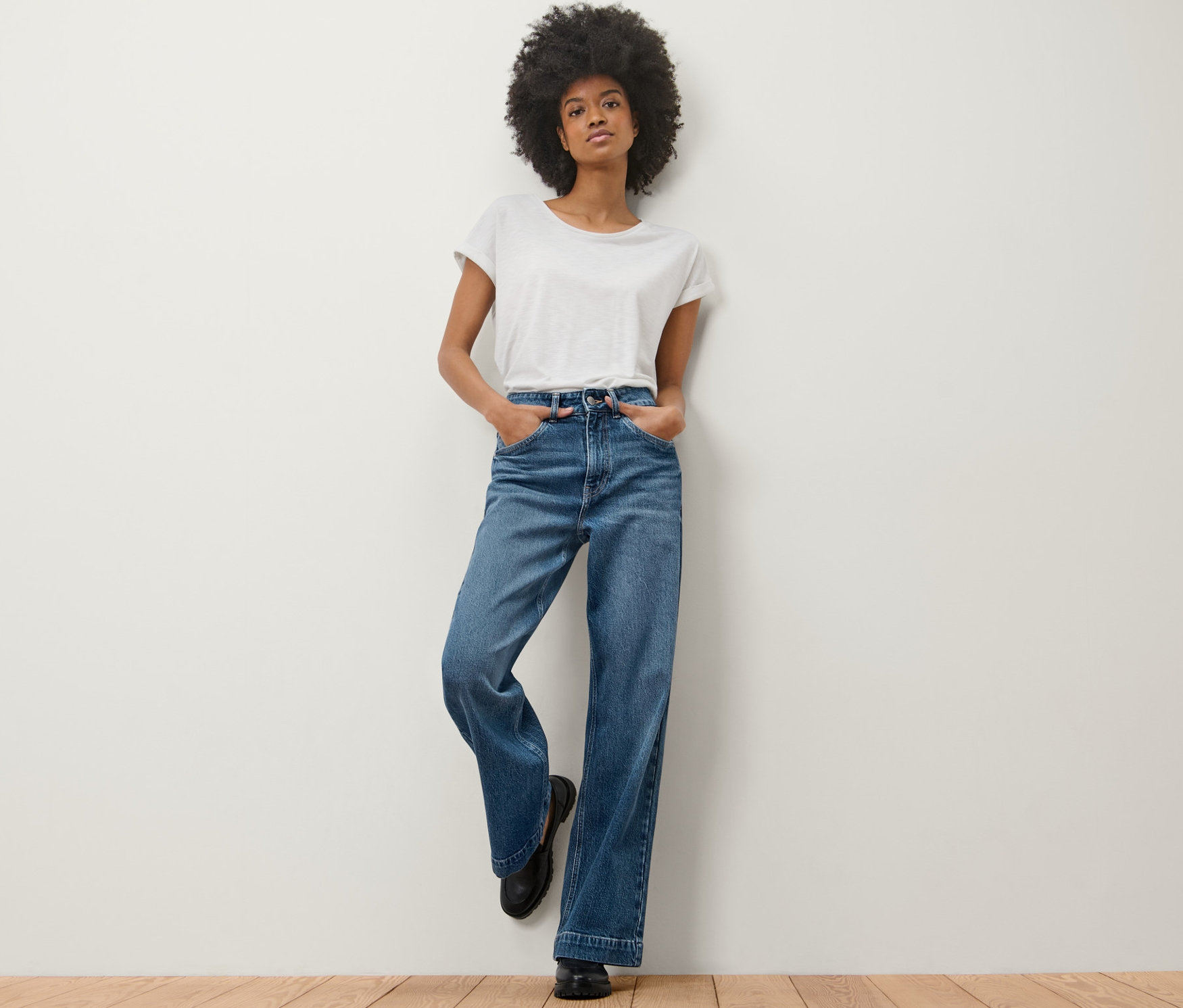 Žena opřená o stěnu má na sobě džíny s širokými nohavicemi – Fit »Viki« a bílé tričko.