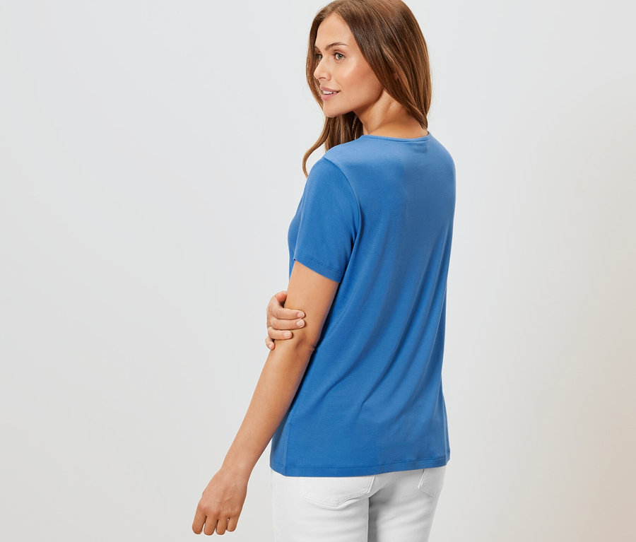 Žena pózuje v kobaltově modrém tričku s detaily skladů a bílých kalhotách.