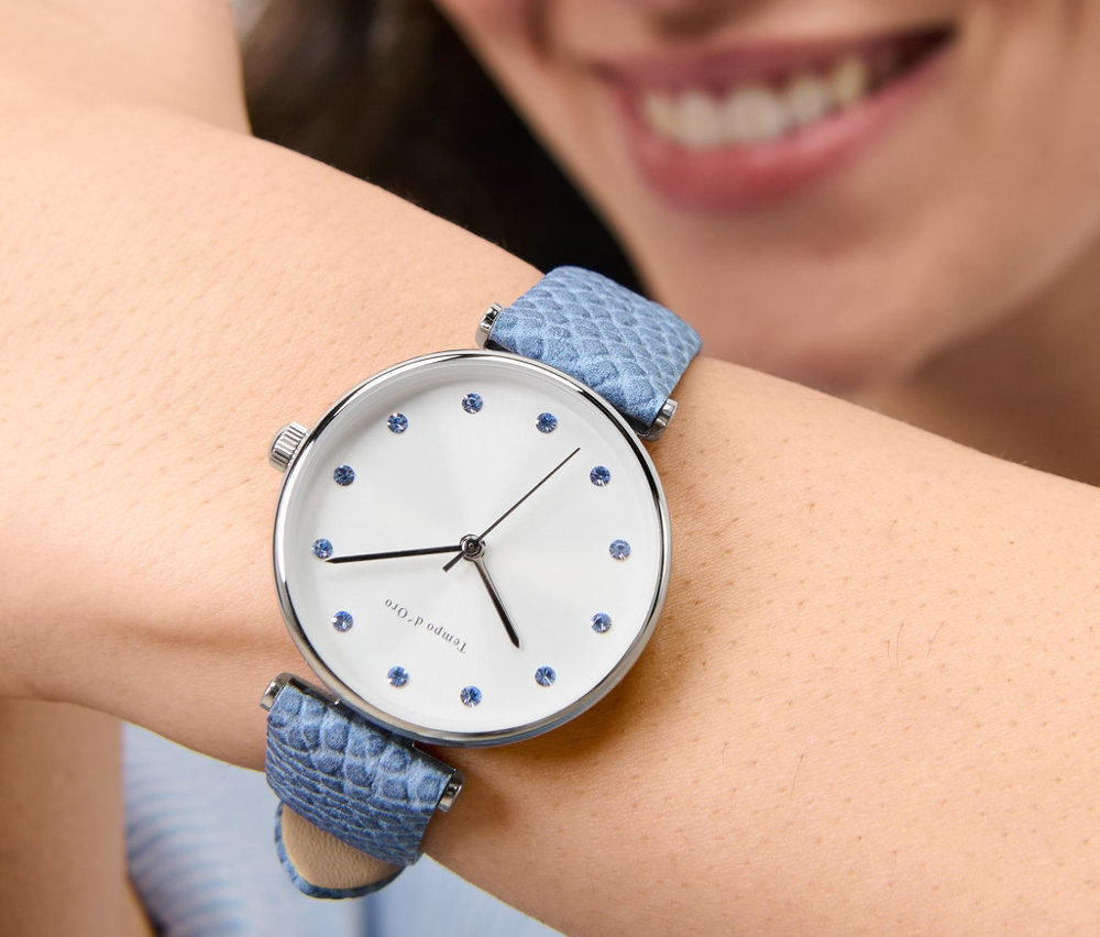 Usmívající se žena má na zápěstí náramkové hodinky s koženým řemínkem a kamínky.