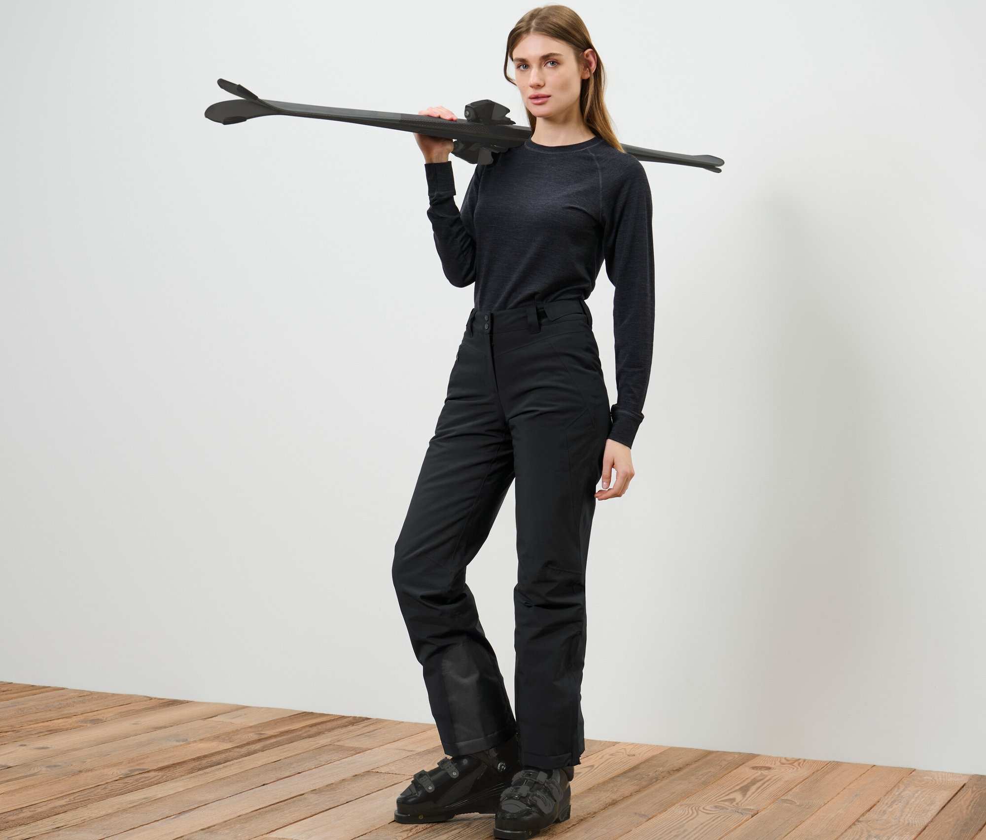 Žena pózuje v lyžařských kalhotách a drží lyže přes rameno.