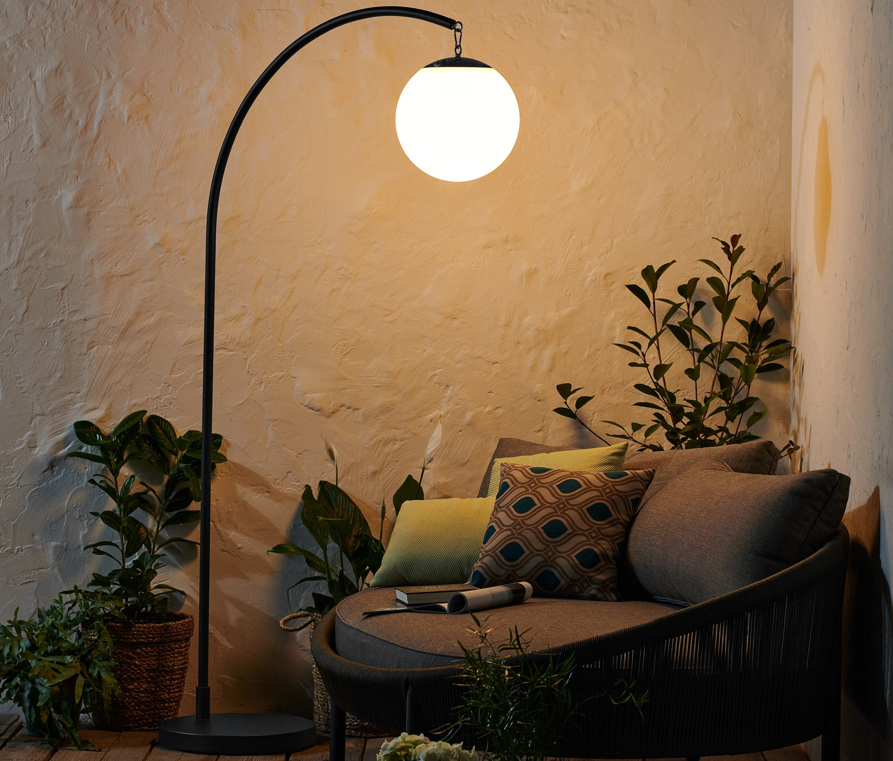 Černá oblouková LED lampa napájená baterií stojí vedle kulaté pohovky s polštáři.