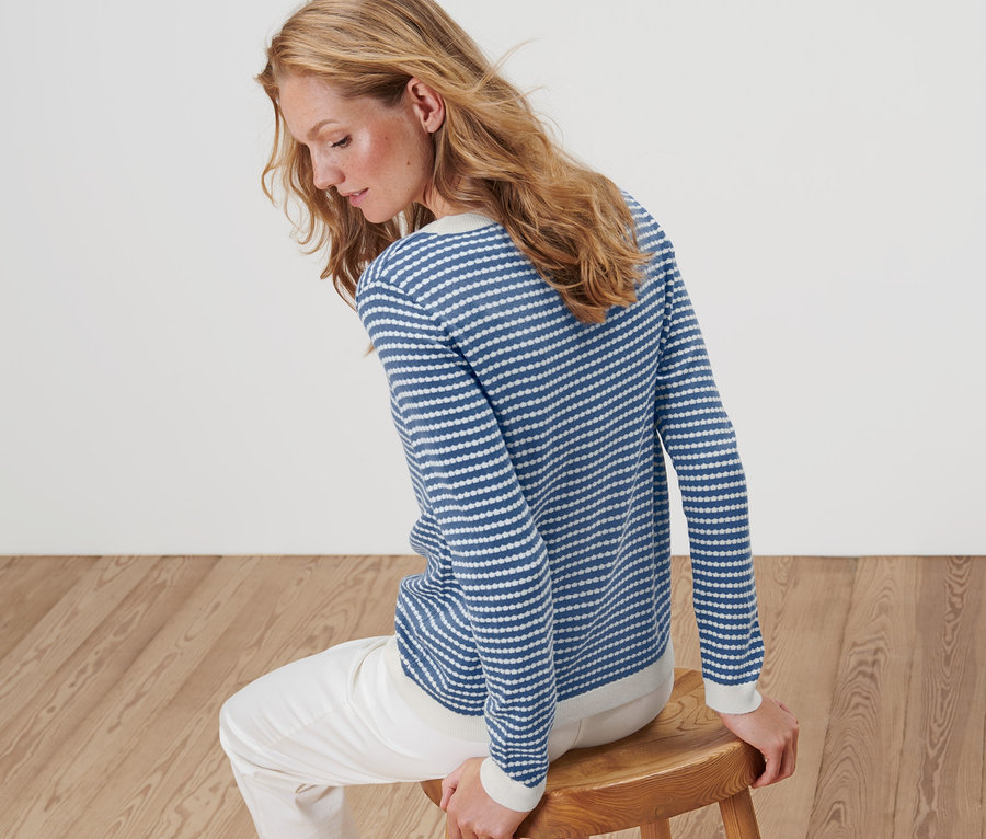 Žena sedí na dřevěné stoličce a má na sobě pruhovaný svetr z jemného úpletu.