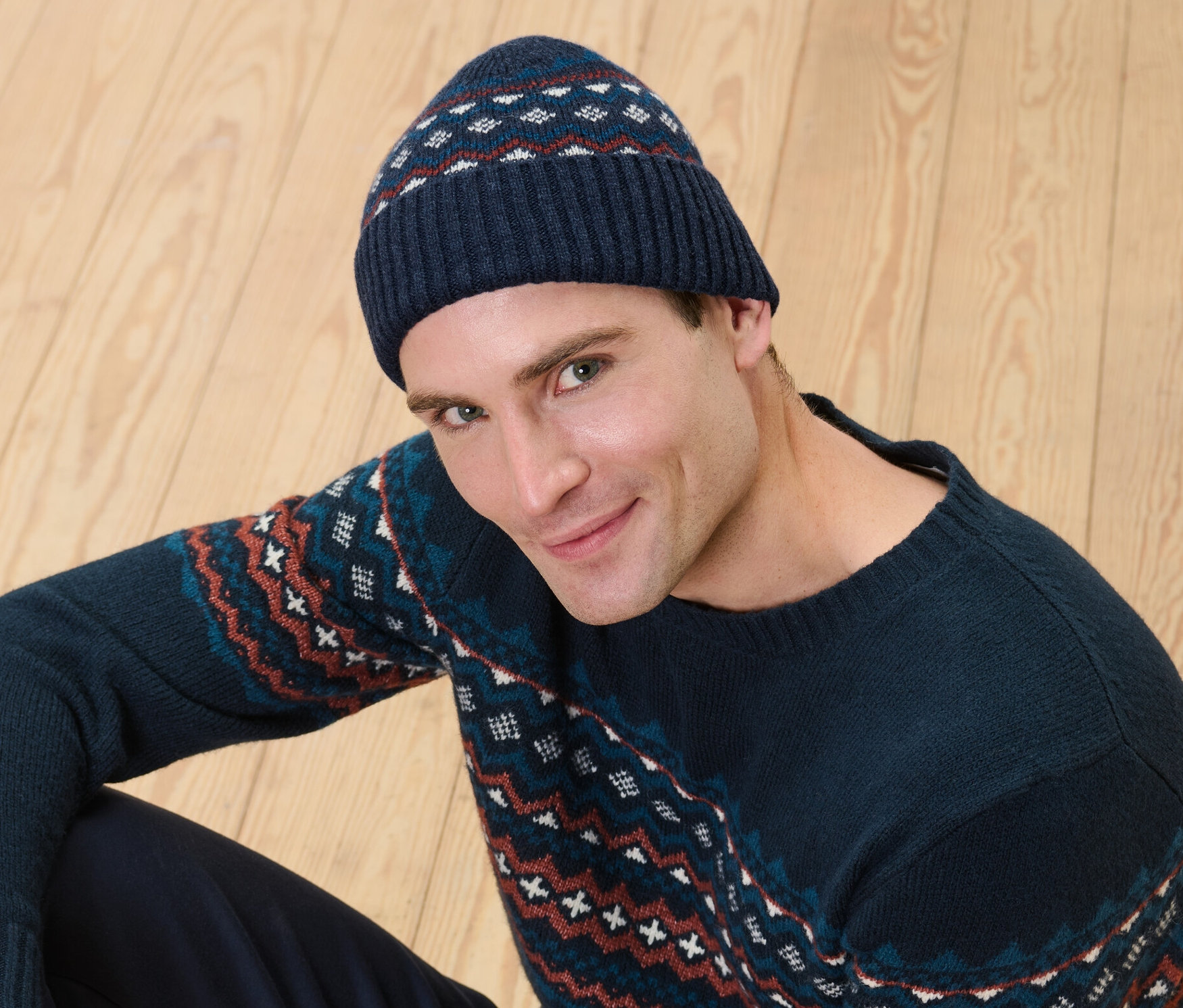 Muž sedí na dřevěné podlaze a má na sobě pletenou čepici s vlnou s certifikací RWS a svetr se vzorem.