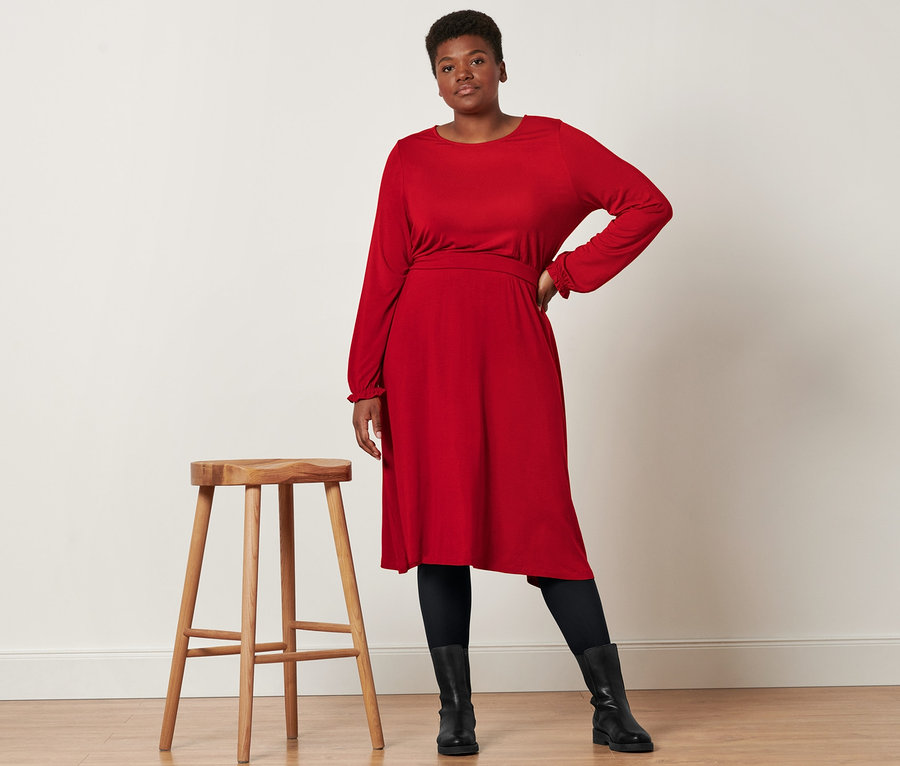 Žena pózuje vedle dřevěné stoličky a má na sobě žerzejové midi šaty, kožené boty a neprůhledné černé punčochové kalhoty (2 páry).