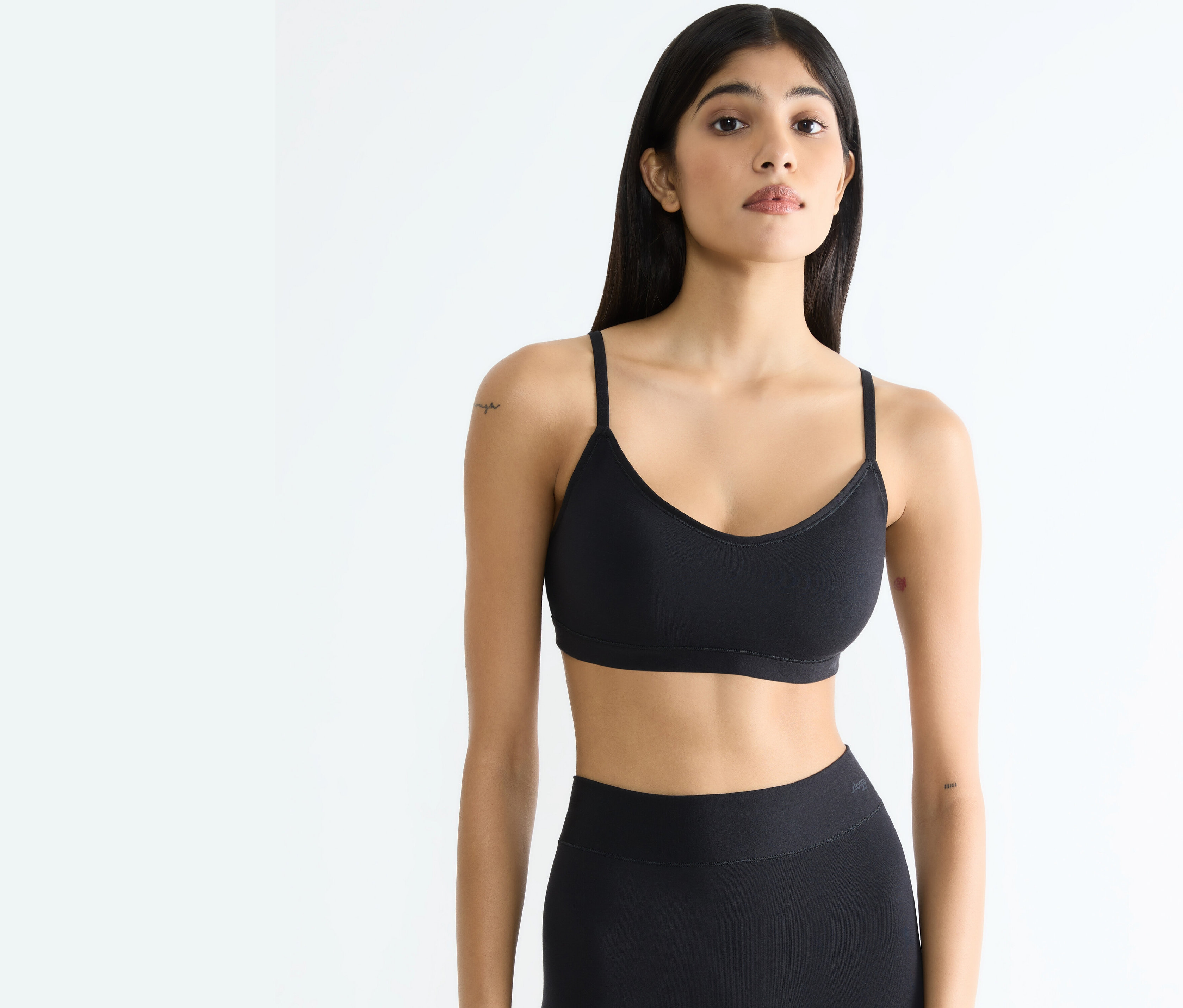 Žena v černém sportovním topu a legínách pózuje na bílém pozadí.