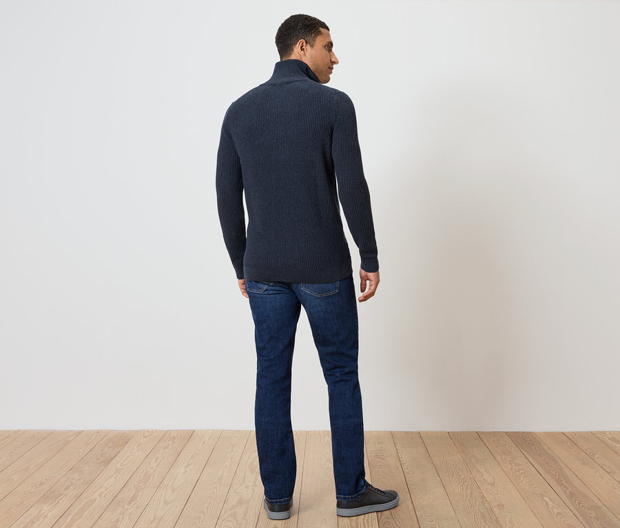 Muž stojí zády ke kameře a má na sobě tmavě modrý svetr a džíny.
