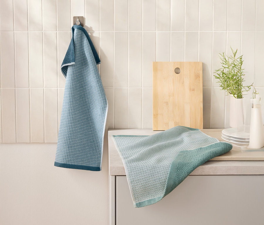 Modrá 2 Waffelpiqué-Küchenhandtücher visí na háku vedle prkénka. Další utěrka leží na kuchyňské lince.