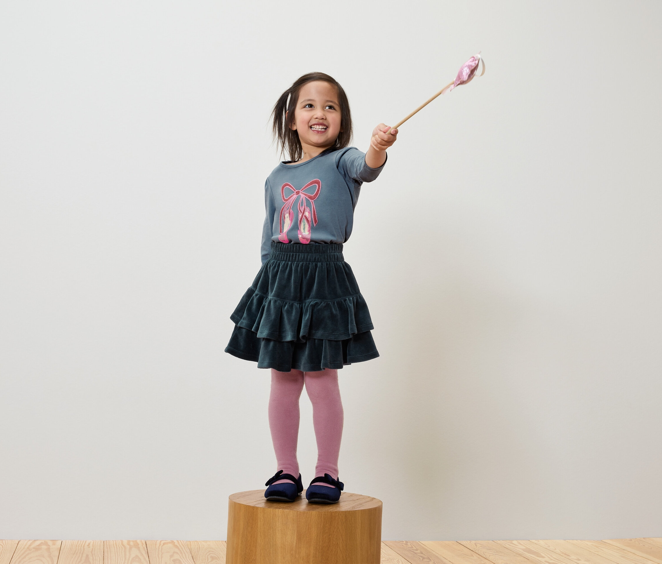 Usmívající se holčička drží kouzelnou hůlku, má na sobě modré tričko s baletními střevíčky, zelenou sukni a růžové punčocháče.