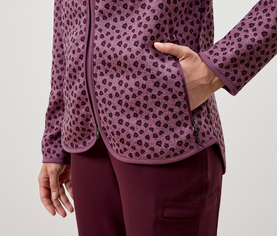 Žena má ruku v kapse fialové bundy se vzorem, oblečené s fialovými kalhotami.