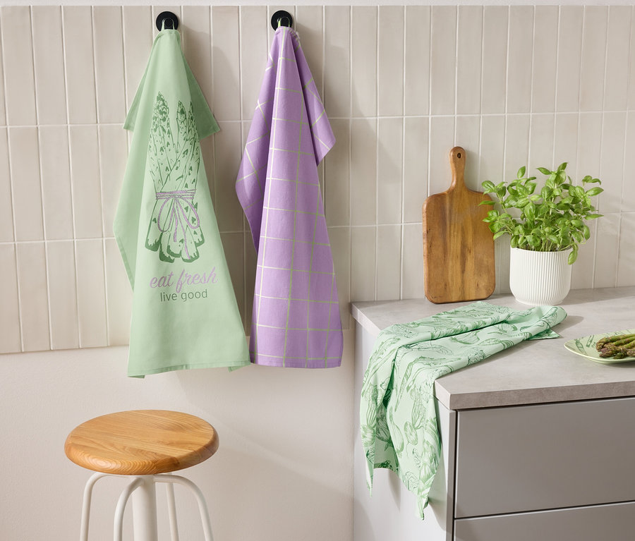 Dvě utěrky visí na zdi vedle sebe, jedna zelená s motivem chřestu a nápisem "eat fresh live good" a druhá fialová s kostkovaným vzorem. Na lince leží zelená utěrka a talíř s chřestem. Vedle stojí prkénko a květináč s bylinkami.