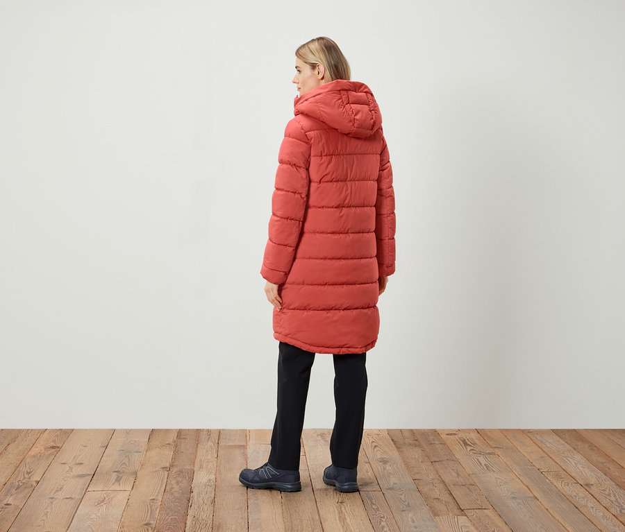 Žena stojí zády, oblečená v červeném prošívaném kabátu s kapucí, černé kalhoty a tmavé boty.