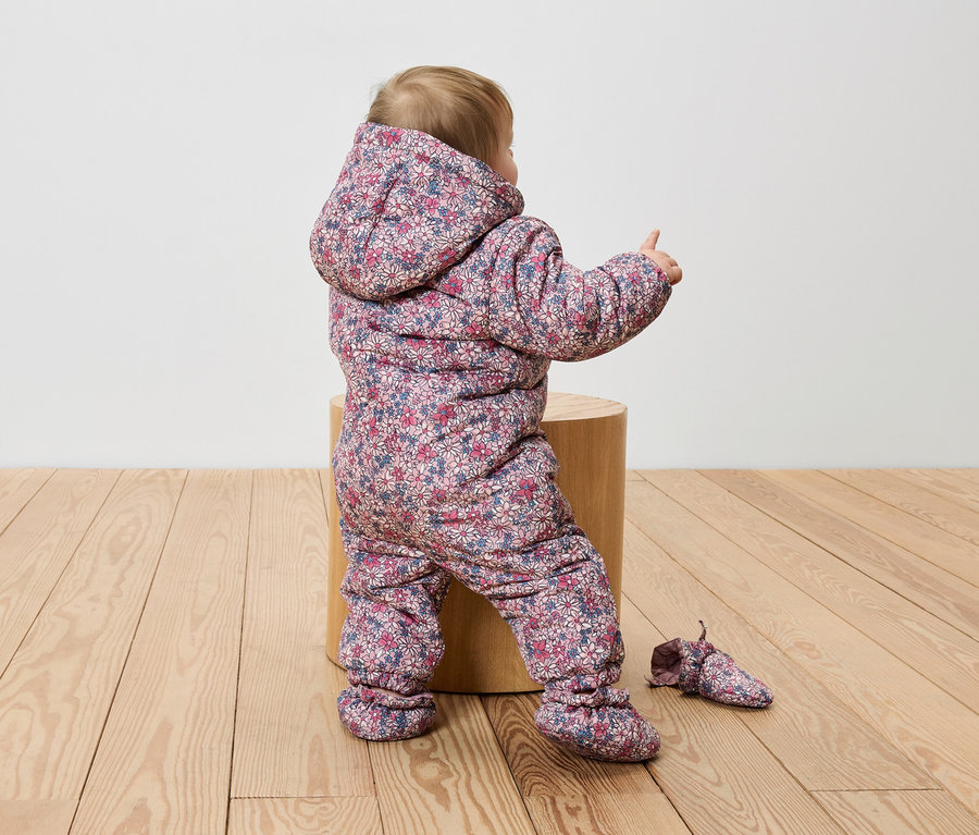 Batole sedí na dřevěném špalku v květinové kombinéze a ukazuje prstem.