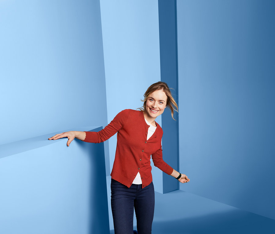 Žena se usmívá a pózuje v červeném kabátku z jemné pleteniny, džínách a bílém tričku na modrém pozadí.