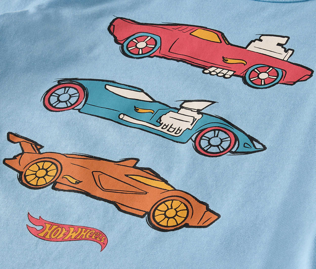 Na světle modrém tričku jsou tři kreslená auta Hot Wheels v červené, modré a oranžové barvě.