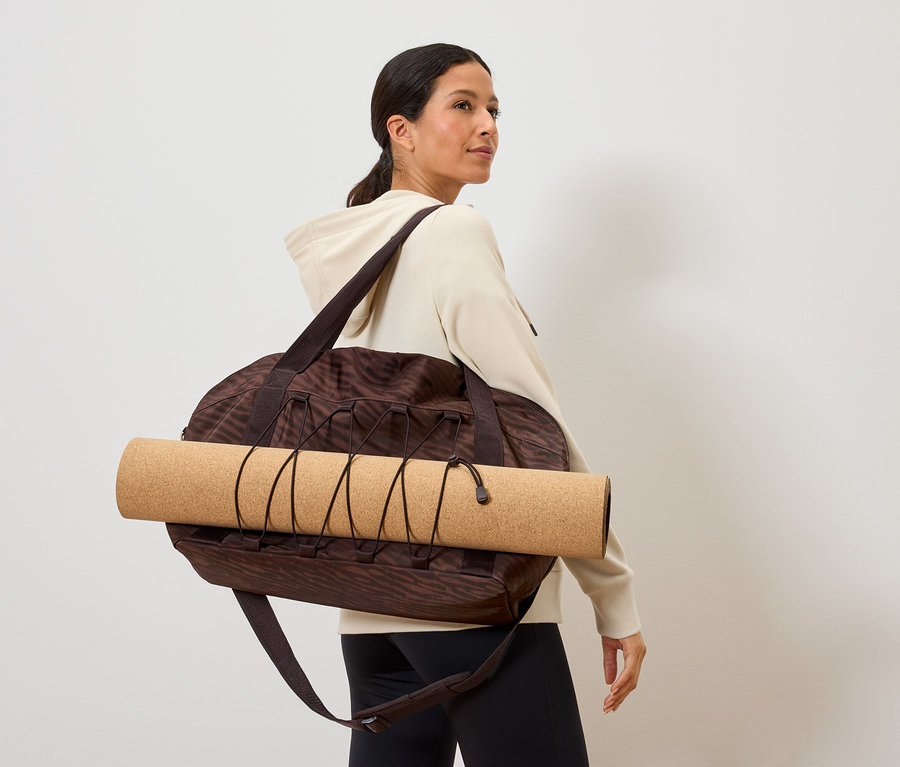 Žena nese hnědou vzorovanou sportovní tašku s podložkou na jógu na ní