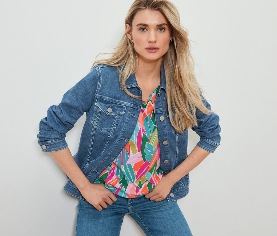 Žena pózuje v džínové bundě, halence se spadlými rameny s celoplošným potiskem a rovných džínách.